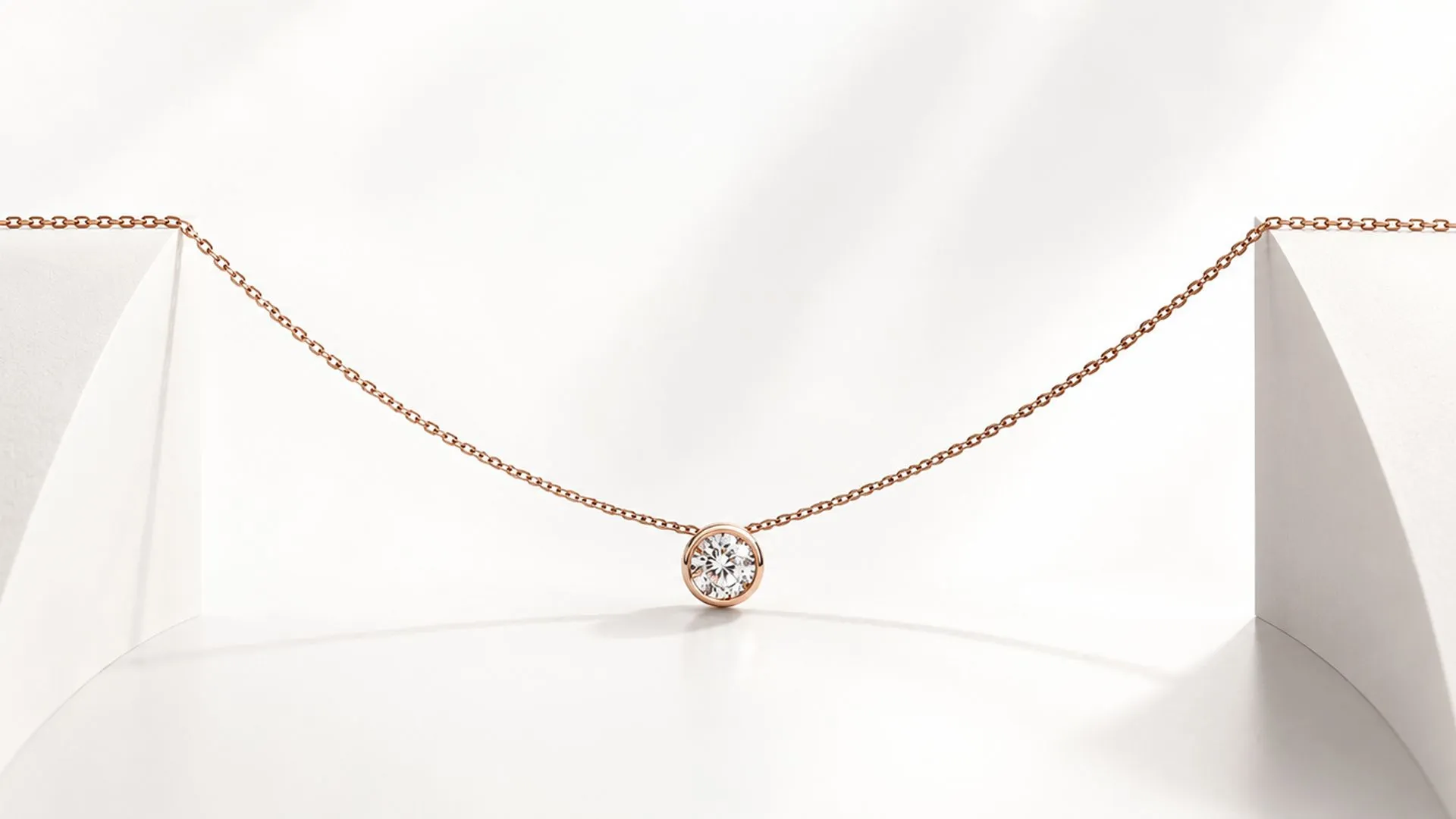 A rose-gold chain necklace with a bezel-set round diamond pendant against a white backdrop